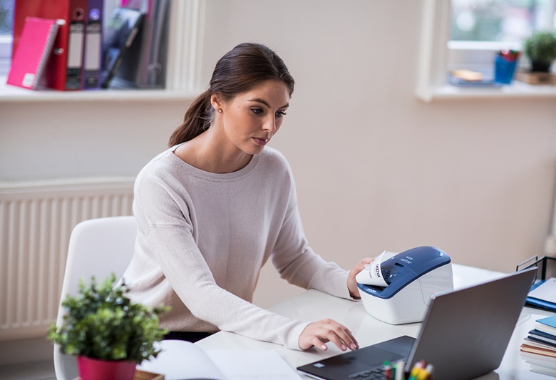 Kobieta siedzi patrząc na laptopa a po jej lewej stronie stoi drukarka drukująca etykietę