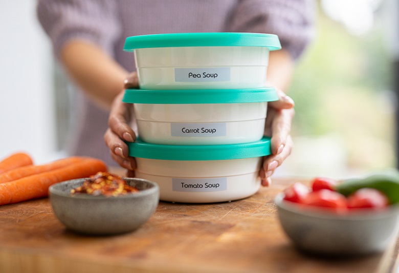 Kobieta trzymająca trzy pudełka Tupperware na desce do krojenia z marchewkami, pomidorami i  ogórkiem