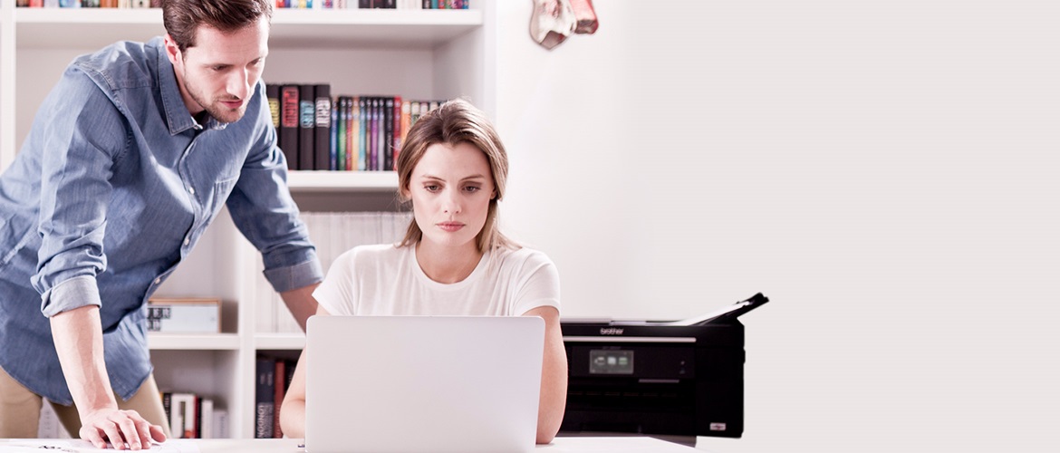 Man and woman collaborating on a laptop and concentrating, with a Brother printer behind them