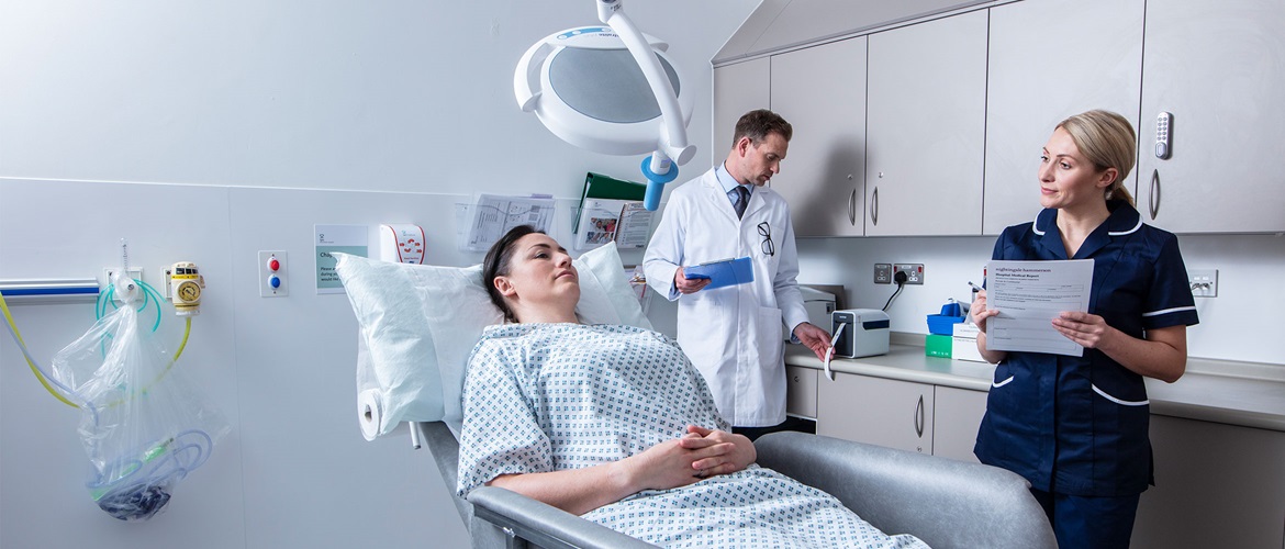 Pacjentka leży na fotelu w pokoju zabiegowym, doktor w białym fartuchu stoi z tyłu, pielęgniarka stoi obok fotela z kartoteką w ręku 